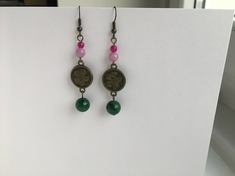 May include: A pair of dangle earrings with a green and pink bead design. The earrings feature a bronze flower charm and a green bead at the bottom.