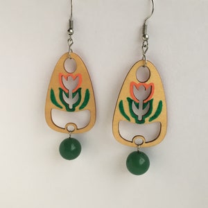 May include: A pair of wooden earrings with a floral design. The earrings are painted in shades of orange, green, and white. Each earring features a green bead dangling from the bottom.