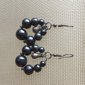 May include: A pair of dark gray beaded earrings. Each earring features a series of dark gray, glossy beads of varying sizes, arranged in a circular shape. The earrings have a hook closure and are displayed against a mesh background.