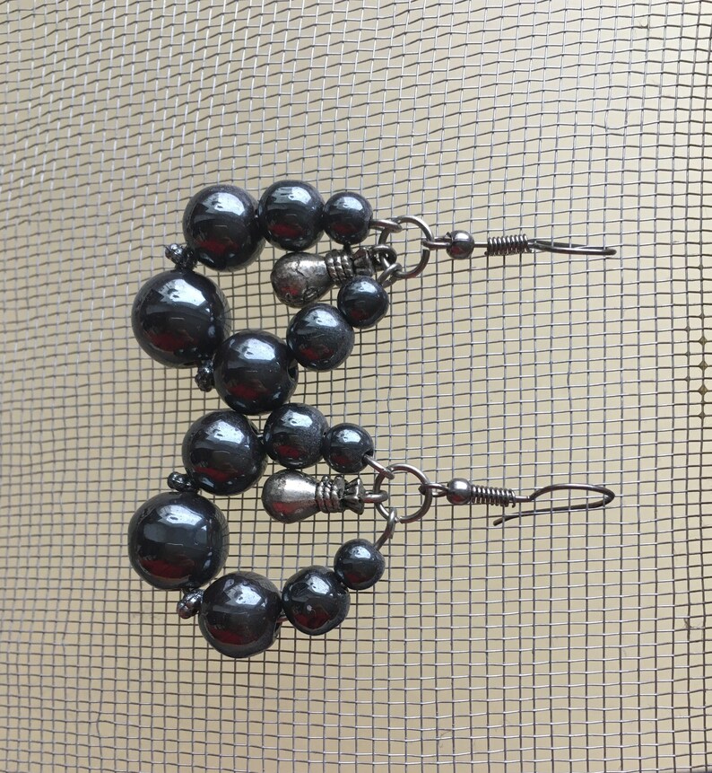 May include: A pair of dark gray beaded earrings. Each earring features a series of dark gray, glossy beads of varying sizes, arranged in a circular shape. The earrings have a hook closure and are displayed against a mesh background.