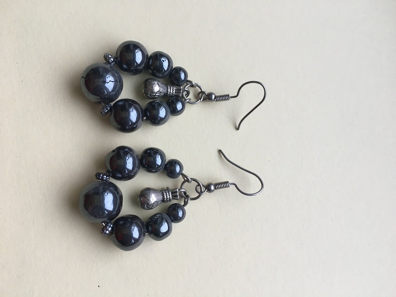 May include: A pair of dark gray beaded earrings. Each earring features a cluster of dark gray beads in varying sizes, connected by silver-toned metal components. The earrings have a hook closure.