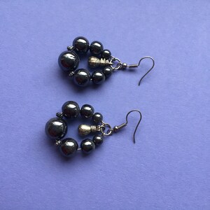 May include: A pair of dark gray beaded earrings. Each earring features a cluster of small beads surrounding a larger bead, with a silver-tone hook. The earrings are displayed on a purple surface.
