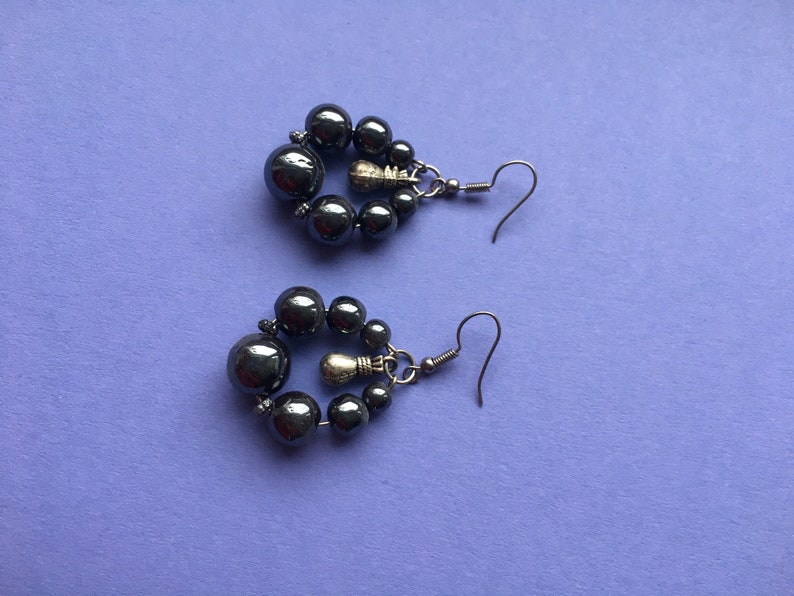 May include: A pair of dark gray beaded earrings. Each earring features a cluster of small beads surrounding a larger bead, with a silver-tone hook. The earrings are displayed on a purple surface.