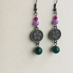 May include: A pair of dangle earrings with a green, purple, and pink bead design. The earrings feature a bronze charm with a flower design.