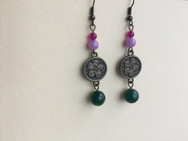 May include: A pair of dangle earrings with a green, purple, and pink bead design. The earrings feature a bronze charm with a flower design.