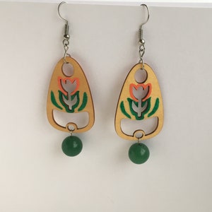 May include: A pair of wooden earrings with a floral design. The earrings are painted in shades of green, orange, and white. Each earring features a green bead at the bottom.