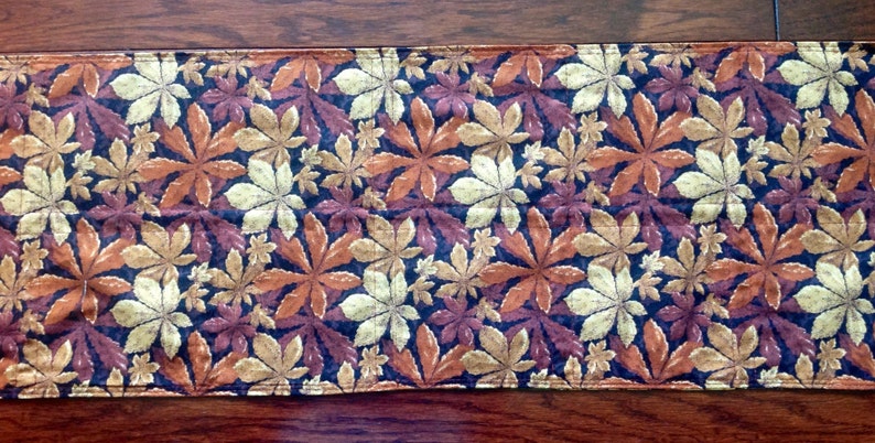 May include: A brown, gold, and cream table runner with a repeating pattern of stylized leaves. The leaves are arranged in a staggered pattern and are outlined in black.