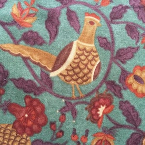 May include: A teal fabric with a repeating pattern of a pheasant, flowers, and foliage. The pheasant is brown, tan, and white with a red crest. The flowers are red and orange, and the leaves are purple.