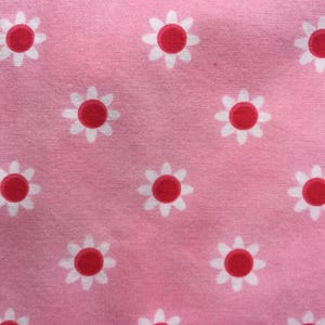 May include: Close-up of a pink fabric with a repeating pattern of white daisies with red centers. The daisies are evenly spaced across the fabric, creating a cheerful and summery design. The fabric has a slightly textured appearance.