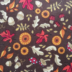 May include: A brown fabric with a repeating pattern of autumn leaves, acorns, mushrooms, and circular designs in shades of red, orange, yellow, and cream. The design is suitable for fall-themed crafts and decor.