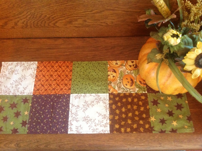 May include: A patchwork table runner with a fall theme. The runner is made of different fabrics in shades of brown, green, orange, and purple. It features a variety of patterns, including stars, leaves, and pumpkins.