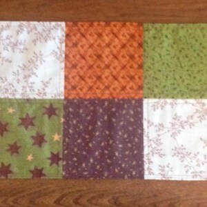 May include: A patchwork table runner with a Halloween theme. The runner is made of various fabrics with different patterns, including pumpkins, stars, and leaves. The colors are brown, orange, green, and white.