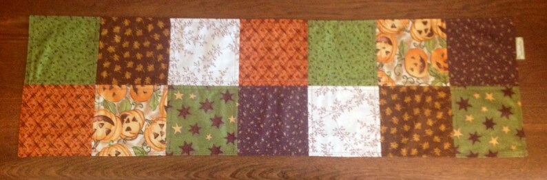 May include: A patchwork table runner with a Halloween theme. The runner is made of various fabrics with different patterns, including pumpkins, stars, and leaves. The colors are brown, orange, green, and white.