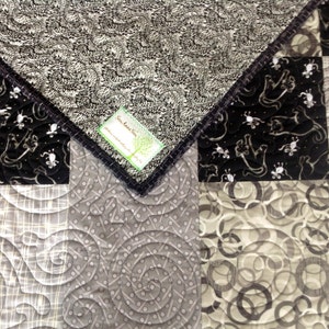 May include: A close-up of a quilt with a patchwork design in shades of black, white, and gray. The quilt features various patterns, including swirls, dots, and a repeating motif of stylized figures. The edges are finished with a dark binding.