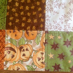 May include: A patchwork quilt with a variety of fall-themed fabrics. The quilt features a brown fabric with a leaf pattern, a white fabric with a brown twig pattern, a green fabric with a brown star pattern, an orange fabric with a crosshatch pattern, and a cream fabric with a pumpkin pattern.