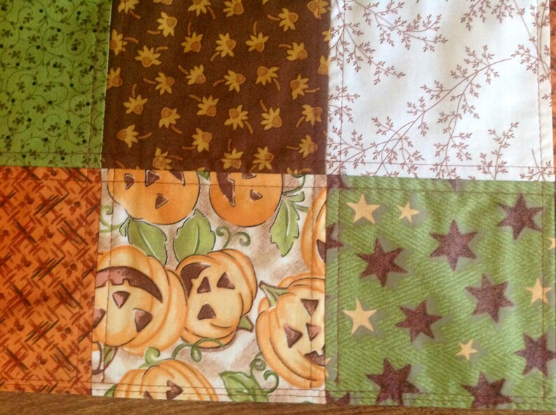 May include: A patchwork quilt with a variety of fall-themed fabrics. The quilt features a brown fabric with a leaf pattern, a white fabric with a brown twig pattern, a green fabric with a brown star pattern, an orange fabric with a crosshatch pattern, and a cream fabric with a pumpkin pattern.