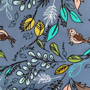 May include: A close-up of a fabric with a blue-gray background featuring a repeating pattern of birds, leaves, and handwritten words. The leaves are in shades of green, yellow, and brown. The words include "beautiful", "happy", and "amazing".