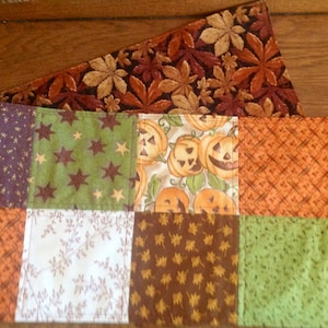 May include: A patchwork table runner with a Halloween theme. The runner is made of different fabrics with various patterns, including pumpkins, stars, leaves, and plaid. The runner is a great way to add a festive touch to your table for the Halloween season.