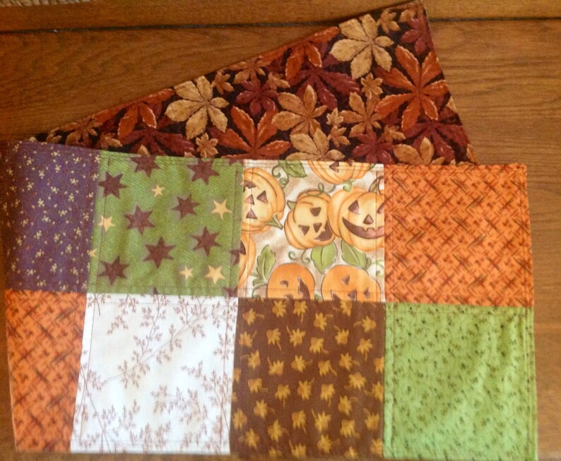 May include: A patchwork table runner with a Halloween theme. The runner is made of different fabrics with various patterns, including pumpkins, stars, leaves, and plaid. The runner is a great way to add a festive touch to your table for the Halloween season.