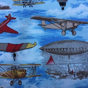 May include: A blue fabric with a repeating pattern of various aircraft, including a red aeroplane, a yellow biplane, a brown airship, and a white glider. The background is a light blue with white clouds.