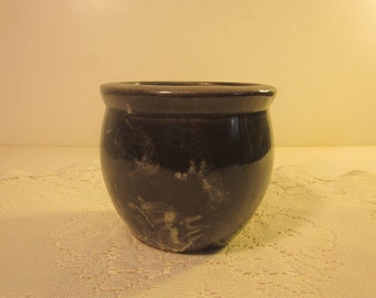May include: A small, dark brown ceramic pot with a rounded body and a slightly flared rim. The pot has a glossy finish with some lighter, textured areas. It is likely used for planting small plants or as a decorative item.