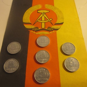 May include: Six silver 5 Pfennig coins are displayed on a flag with black, red, and yellow stripes, and a central emblem. The coins are arranged in a pattern, with the number 5 and the word "Pfennig" visible.