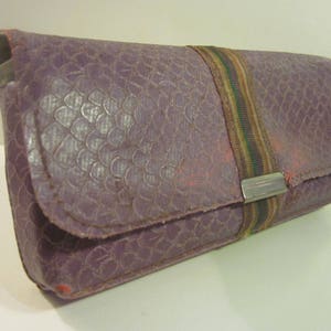 May include: A purple leather clutch with a textured pattern and a brown and green striped strap. The clutch has a silver metal clasp.