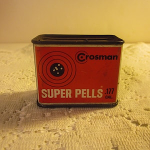 May include: A rectangular red tin with black trim, featuring the Crosman logo and the words "SUPER PELLS .177 CAL." A target graphic is on the front. The tin appears to be for air gun pellets.