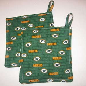 Könnte beinhalten: Zwei grüne und goldene Topflappen mit dem Wort "Packers" darauf. Die Topflappen haben einen Schlaufe.