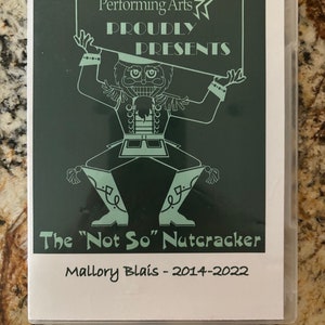 Può includere: Un'illustrazione verde e bianca di uno schiaccianoci che tiene un cartello con su scritto "Center for the Performing Arts Proudly Presents The "Not So" Nutcracker Mallory Blais - 2014-2022".