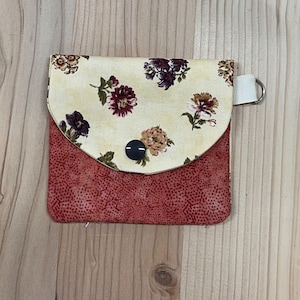 May include: A small, square fabric wallet with a floral pattern on a cream-colored flap and a red dotted base. The flap is secured with a black button. A white loop with a silver ring is attached to the side.