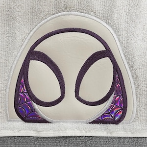 May include: A white and gray towel featuring an embroidered design of a superhero mask. The mask has a white background with purple accents and a holographic pattern. The towel is made of a soft, absorbent material.