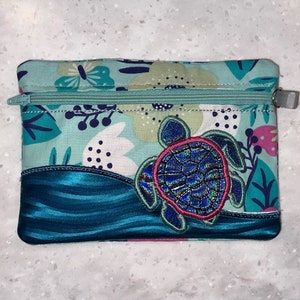 May include: A small, rectangular zippered pouch with a teal floral pattern and a blue wave design. A blue and pink sea turtle applique is centered on the pouch. The zipper is teal with a silver pull.
