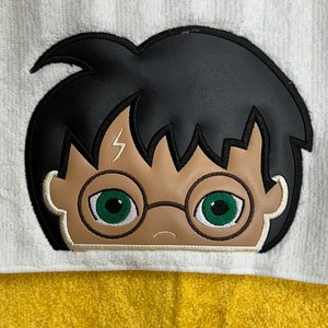 Op de afbeelding: Een witte badjas met capuchon met een geborduurd zwart en bruin beeld van Harry Potters gezicht. De badjas heeft een gele badstof voering.