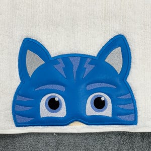 Puede incluir: Una cara de gato azul y gris bordada en una toalla blanca. El gato tiene ojos negros y una máscara azul.