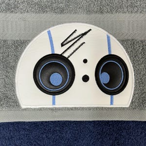 May include: A white towel with a blue and black embroidered robot face on it. The robot has two large black eyes with blue pupils and a black mouth.