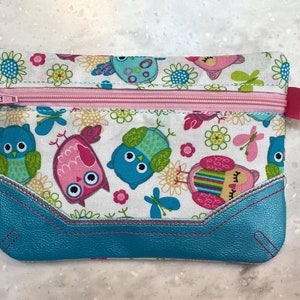 May include: A rectangular zippered pouch featuring a whimsical owl pattern in shades of blue, pink, and green on a white background. The pouch has a turquoise bottom with pink stitching and a pink zipper. A pink ribbon is attached to the zipper.