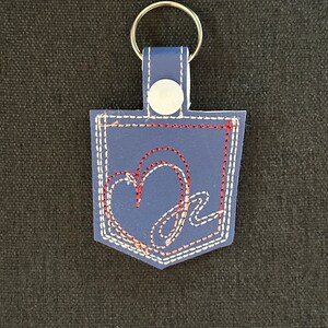 Peut inclure: Un porte-clés en cuir bleu avec un motif de poche cousu en blanc. La poche a un cœur brodé rouge et blanc avec un "M" en cursive à l'intérieur.