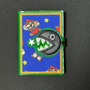 Może przedstawiać: Niebieski portfel z tkaniny z zieloną obwódką i czarno-białą postacią z serii gier wideo Super Mario Bros. Postać to duża, okrągła, czarna istota z jednym okiem i ostrymi zębami. Portfel ma zamek błyskawiczny i małą kieszeń z przodu.