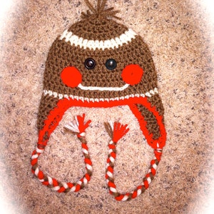 May include: A handmade brown crochet hat with white stripes, black button eyes, and red cheeks. The hat has red earflaps and braided ties with white and red accents. A whimsical, cozy winter accessory.