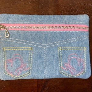 Könnte beinhalten: Eine Jeans-Optik-Tasche mit einem rosa Reißverschluss und einem silbernen Anhänger. Die hellblaue Tasche hat gestickte rosa herzförmige Designs auf der Vorderseite, die wie Jeanstaschen aussehen. Weiße Nähte detaillieren das Design.