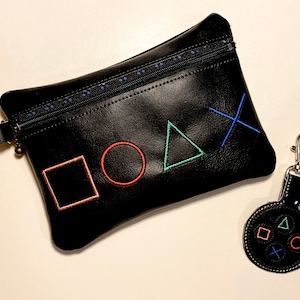 May include: Black zippered pouch and matching keychain featuring embroidered gaming controller symbols in red, green, blue, and orange. The pouch has a wrist strap and a blue dotted zipper.