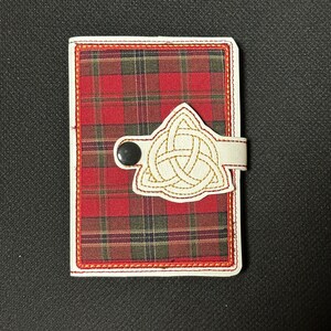 May include: A rectangular wallet featuring a red, green, and black tartan pattern. It has a white border and a white strap with a gold Celtic knot design. A black button secures the strap.