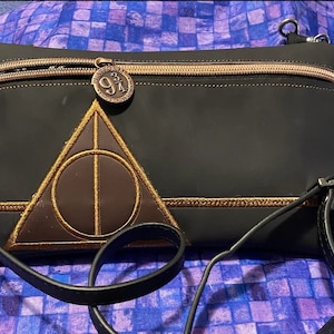 May include: Black crossbody bag with a gold zipper and a brown and gold Deathly Hallows symbol. The bag has a Platform 9 3/4 charm and a black strap. The bag is made of a shiny, smooth material.
