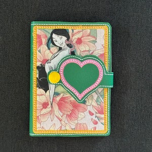May include: Green and floral patterned wallet featuring a Mulan illustration. The wallet has a green heart-shaped clasp with pink stitching and a yellow button closure. The edges are trimmed with yellow stitching.