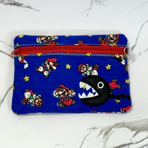 Könnte beinhalten: Blaue Reißverschlusstasche mit einem sich wiederholenden Muster von Mario-Charakteren und gelben Sternen. Auf der Vorderseite ist ein schwarzer Chain Chomp-Charakter aufgestickt. Der Reißverschluss ist rot.