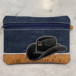 May include: Denim zippered pouch with a black cowboy hat applique. The hat has the initials "HR" embroidered on it. The bottom of the pouch is made of cork material.