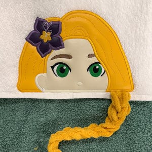 以下が含まれることがあります： 白いタオルに、漫画のキャラクターの顔の刺繍が施されています。キャラクターは金色の髪、緑色の目、紫色の花を持っています。長い金色の三つ編みが垂れ下がっています。タオルの下部は緑色です。