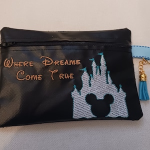 Puede incluir: Bolsa negra con cremallera con una imagen bordada del castillo de Cenicienta y Mickey Mouse. El texto "Where Dreams Come True" está bordado en hilo dorado.