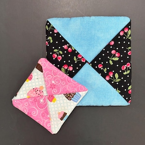 May include: Two fabric coasters. One is square with a light blue center and black corners with white polka dots and red cherries. The other is a smaller, folded square with pink and white cupcake patterns.